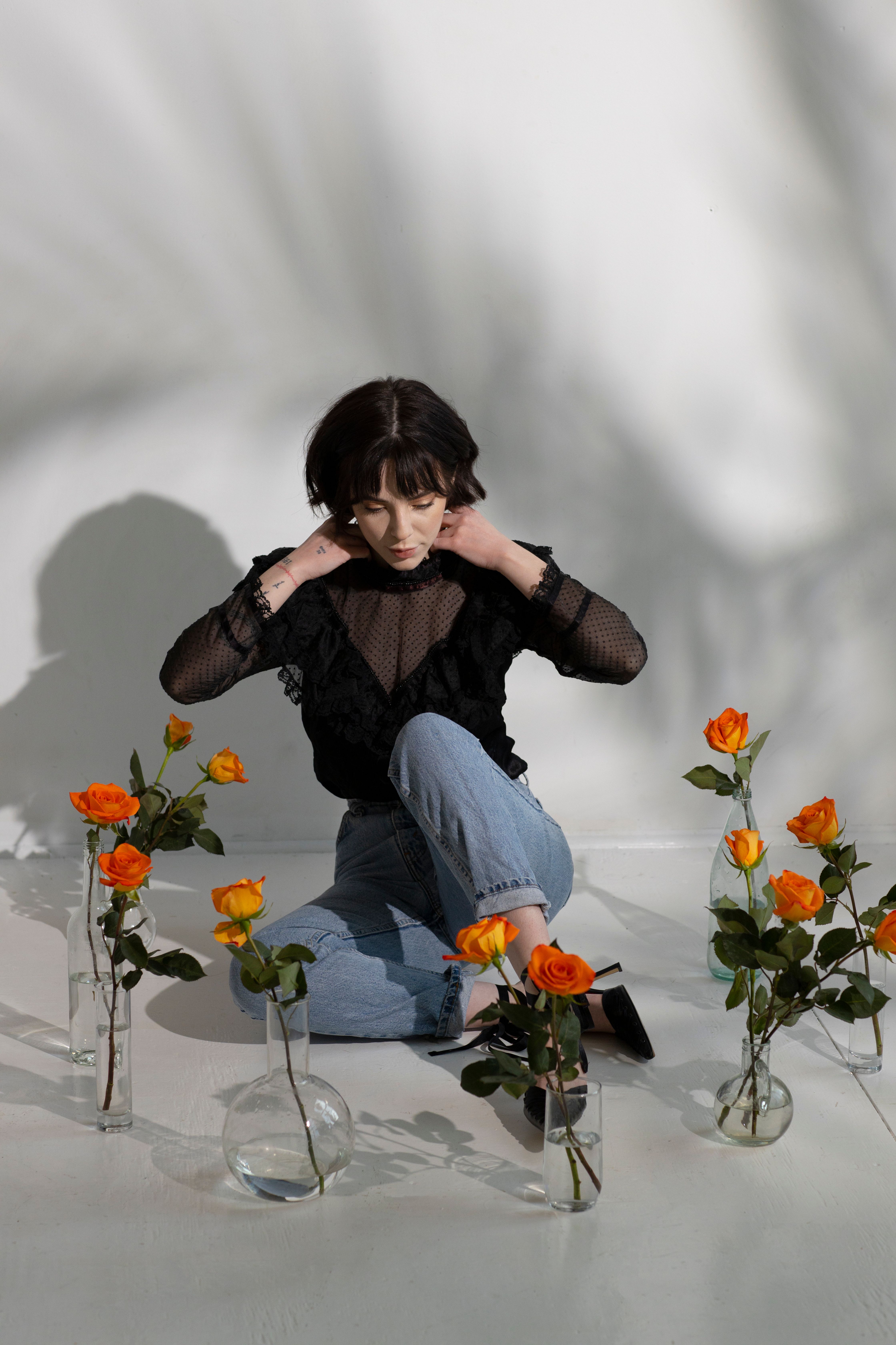 Full body of young thoughtful female in trendy clothes sitting on white floor with glass vases with flowers near white wall with shadows