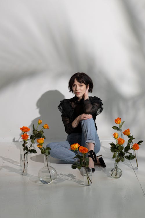 Full length of young pensive female in stylish outfit sitting on tile floor surrounded with glass vases with orange roses near white wall with shades while looking at camera