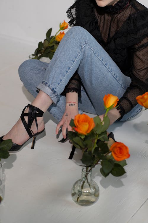 Crop faceless female in stylish outfit and high heels sitting on tile floor with shades near glass vases with orange roses in light studio