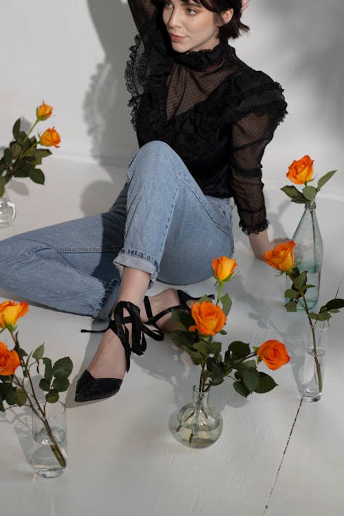 Young thoughtful female in trendy clothes sitting on floor while looking away near orange roses in glass vases near white wall with shadows