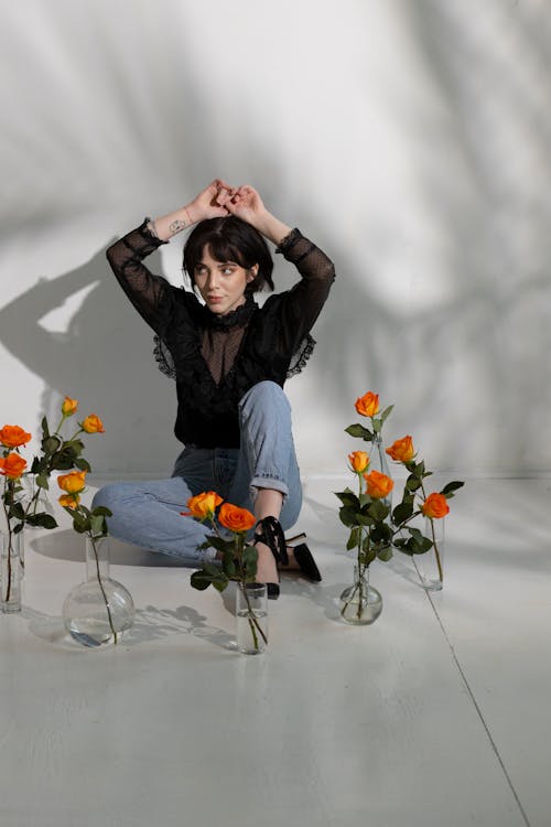 Full body of young dreamy female in trendy clothes sitting on white floor with glass vases with roses near white wall with shadows while looking away with raised hands