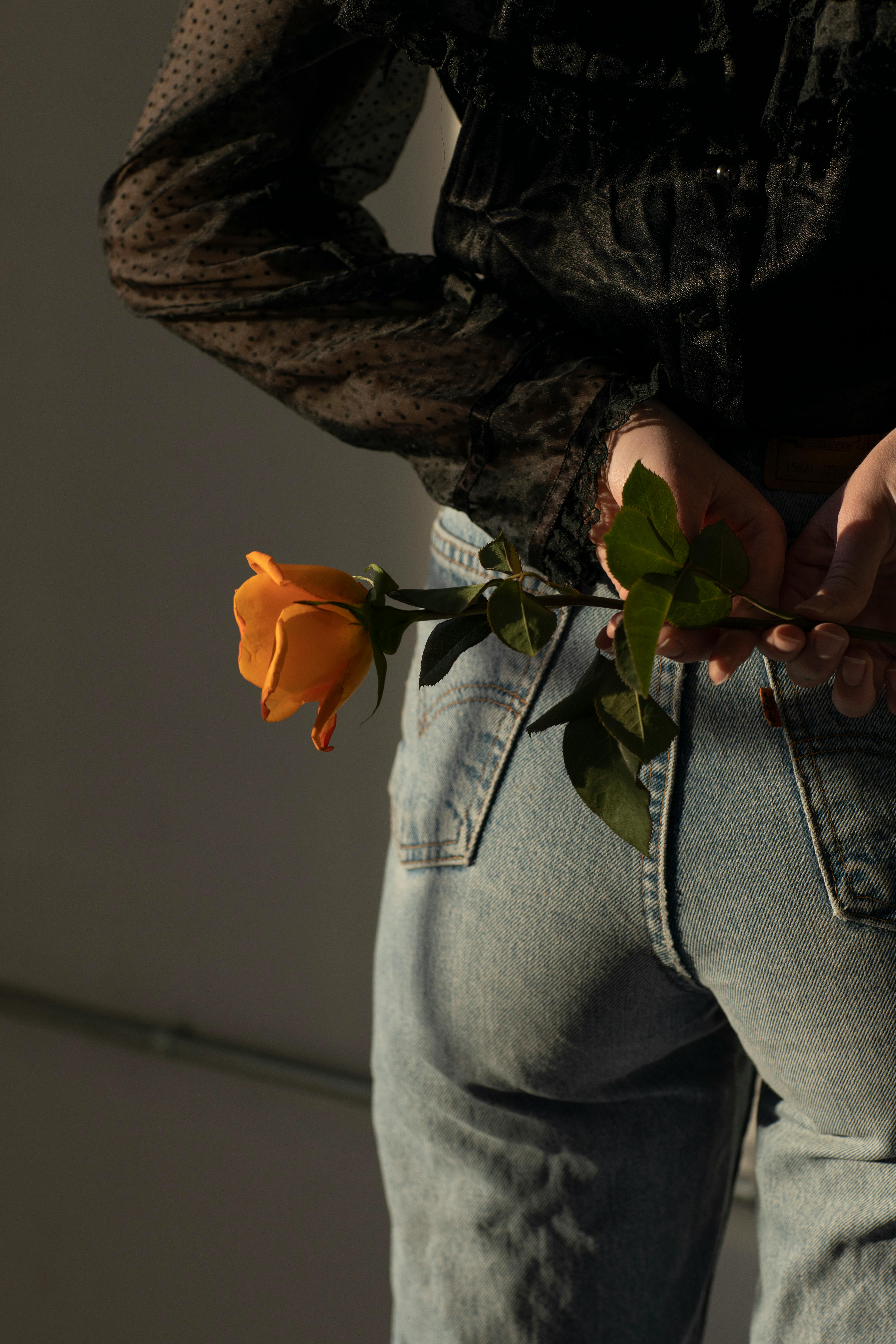Close-up of a woman holding an orange rose behind her back, wearing denim jeans.