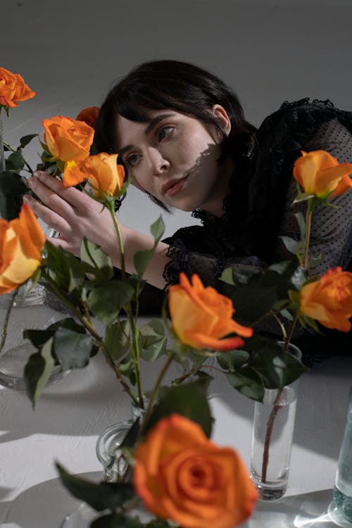 Free Young gothic female with short dark hair looking at tender orange roses with fresh green stems on gray background Stock Photo