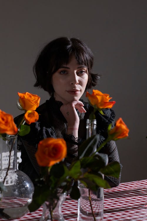 Gentle woman touching chin at table with orange delicate roses