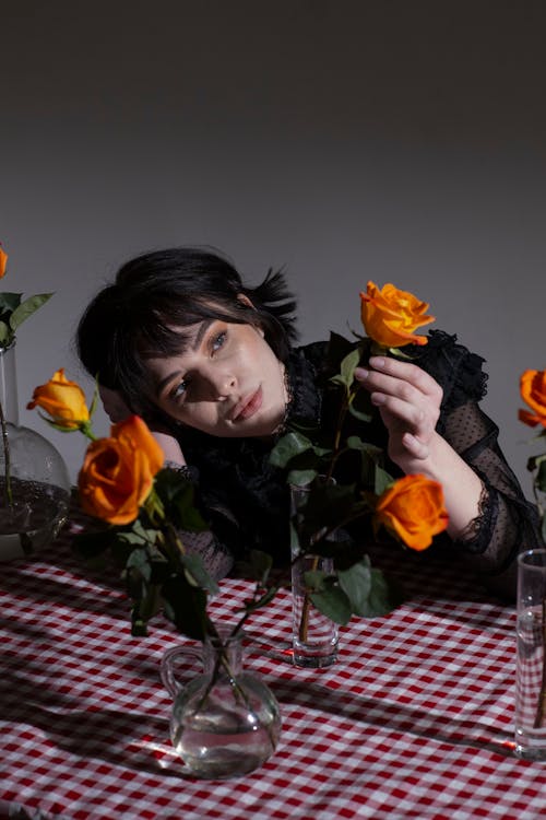 Elegant female with dark short hair touching green stems of orange blooming roses at table