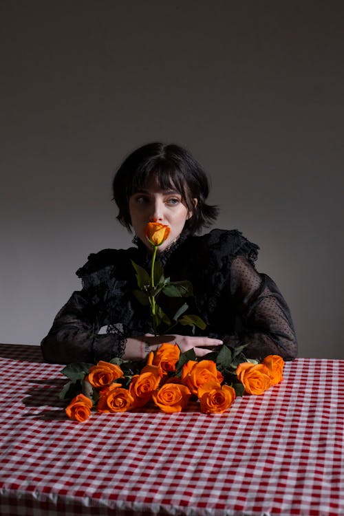 Thoughtful female with fragrant blooming orange rose looking away at table with bouquet on gray background