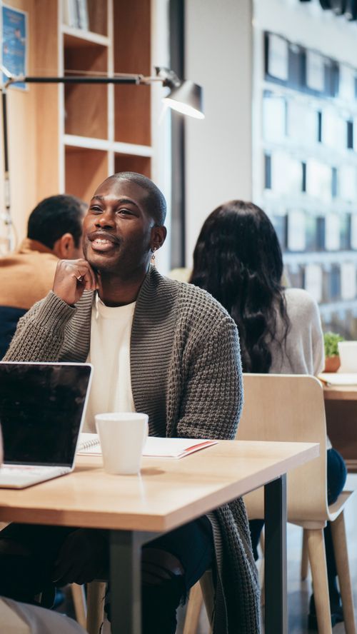 Free Man smiling while sitting at a table with a laptop and coffee in a cozy indoor setting. Stock Photo