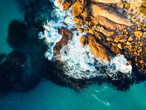 Free A breathtaking aerial shot of waves crashing on a rocky shoreline with vibrant blue water. Stock Photo