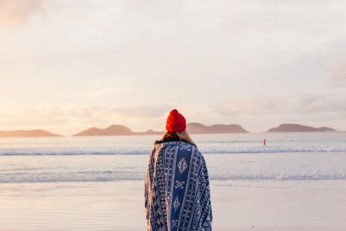 Free A woman wrapped in a blanket stands on a serene beach at sunset. Stock Photo