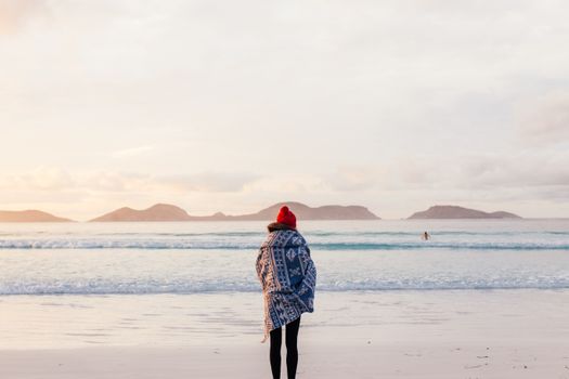 Woman in a blue patterned poncho and red hat standing on a tranquil beach at sunset.