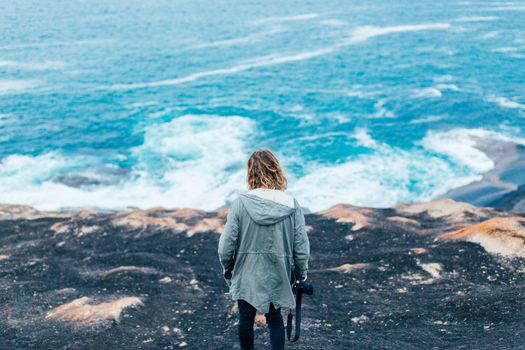 A woman holding a camera stands on a rocky cliff, overlooking the vast ocean.