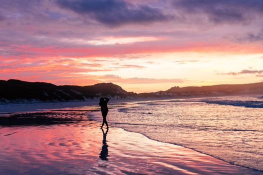 A person stands silhouetted on a beach against a stunning, colorful sunset with calm waves.