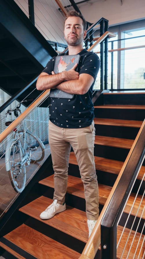Free Man in Black Crew Neck T-shirt and Brown Denim Jeans Standing on Brown Wooden Staircase Stock Photo