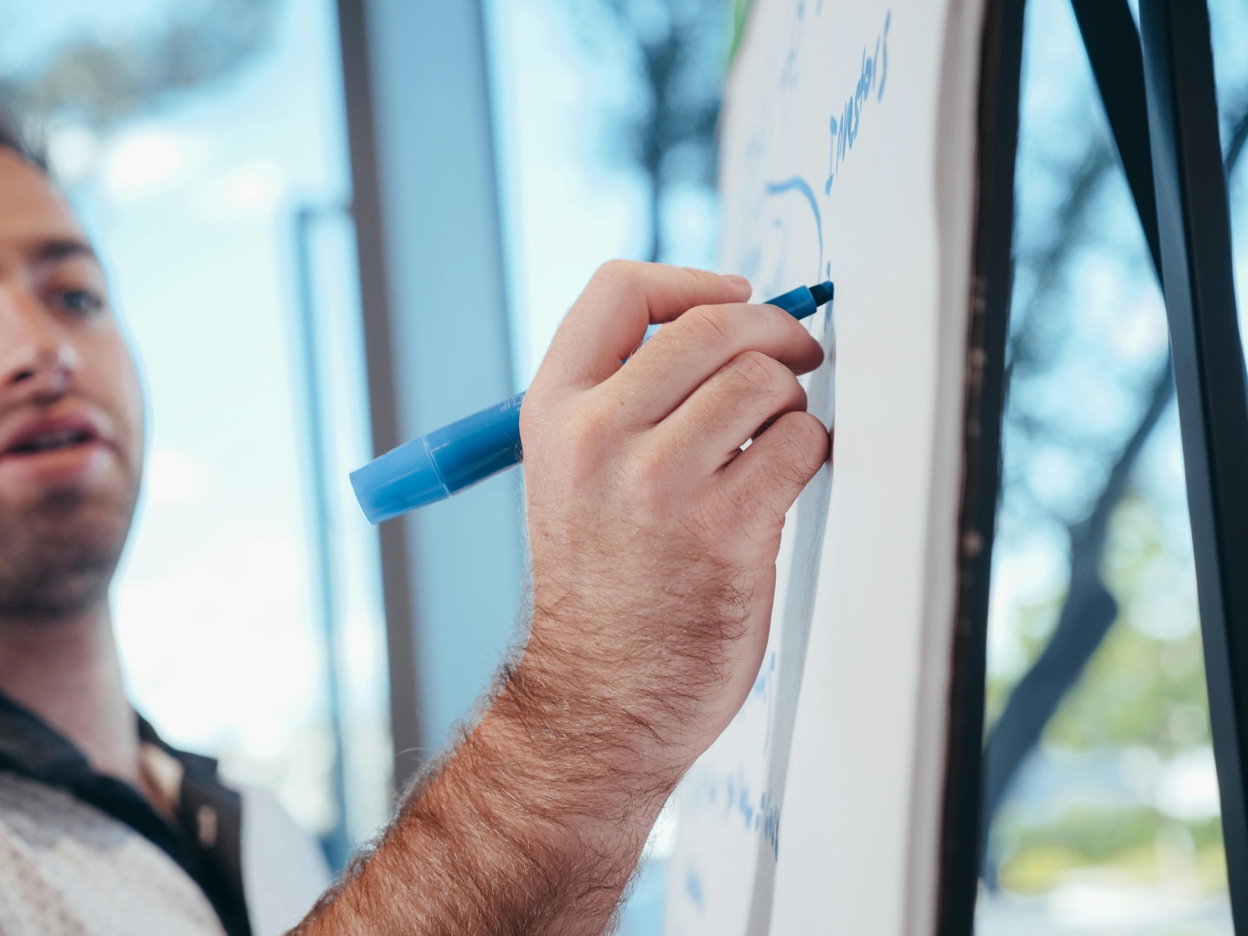 A person writing with a blue marker on a whiteboard, close-up view in an office setting.
