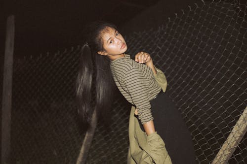 Side view of serious ethnic lady in trendy clothes standing near metal grid at night in street and looking over shoulder