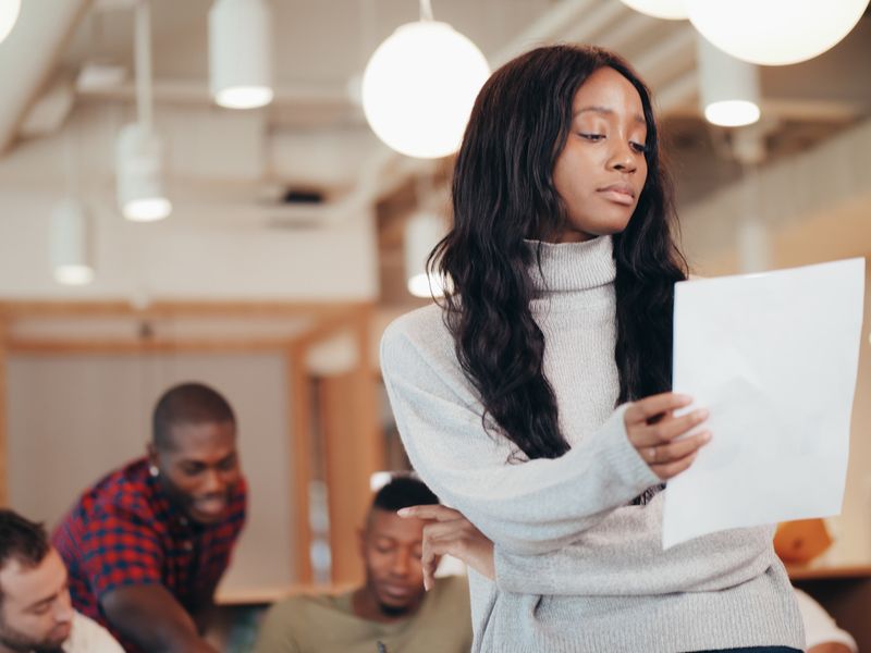 A young Black woman in a gray turtleneck reads a sheet of paper in an office, with coworkers out of focus behind her.
