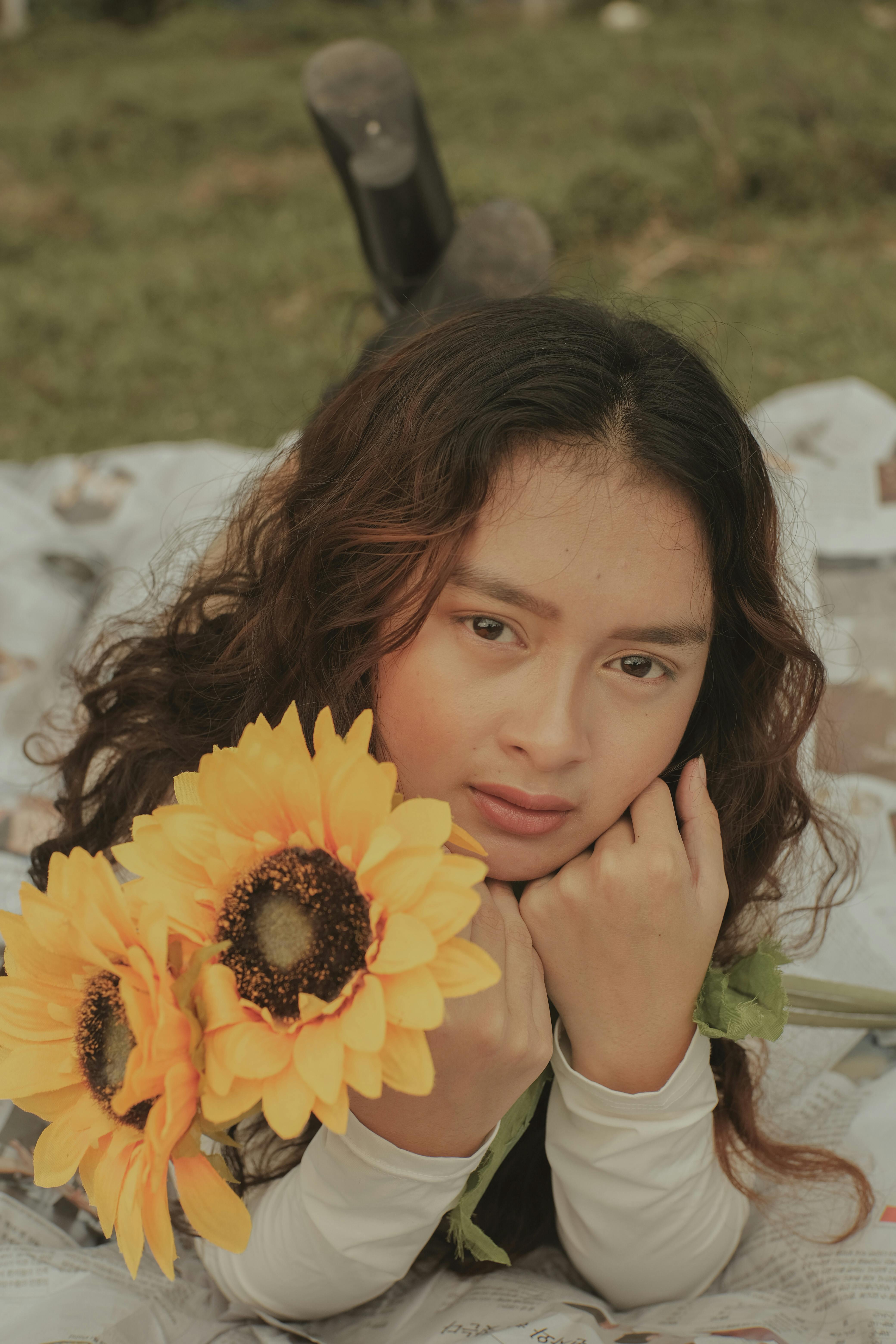 Thoughtful young Asian lady with yellow sunflowers in hand looking at camera while lying on newspapers on grassy green field in daylight in nature