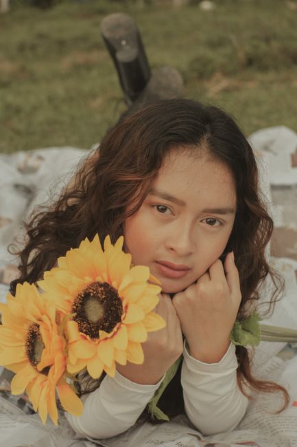 Thoughtful young Asian lady with yellow sunflowers in hand looking at camera while lying on newspapers on grassy green field in daylight in nature