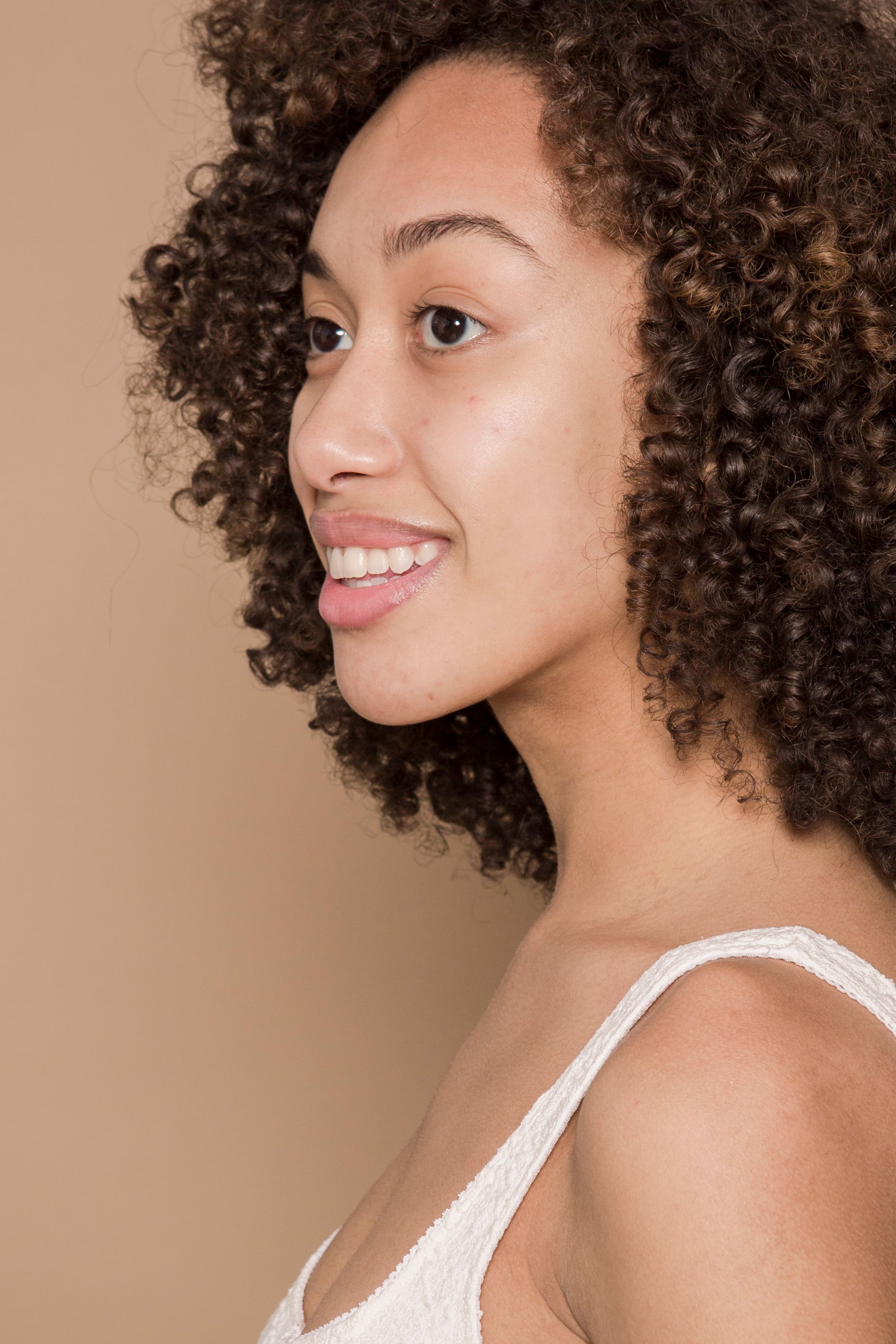 Side view of positive ethnic female with dark hair wearing white outfit smiling and looking away against beige background in studio