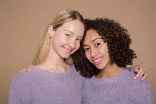 Cheerful multiracial female friends with graphic eyeliner wearing similar clothes hugging and looking at camera while standing against beige background