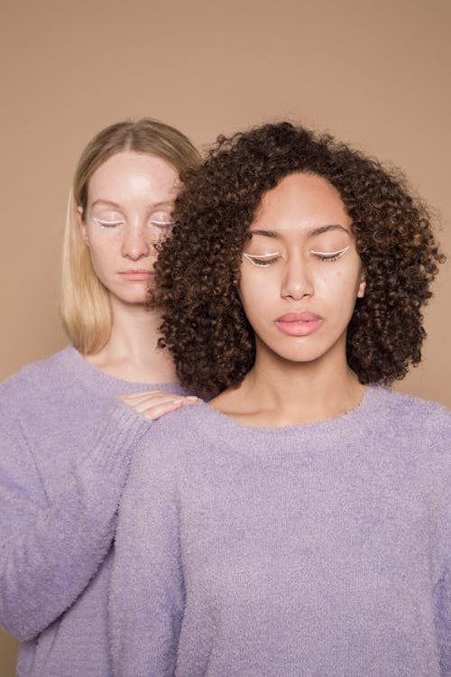 Tranquil multiracial female friends with closed eyes and colorful graphic eyeliner wearing purple sweaters standing against beige background in studio