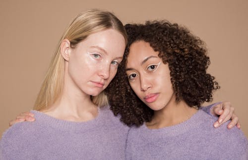 Free Calm multiracial female friends with colorful makeup wearing similar clothes embracing and looking at camera against beige background in studio Stock Photo