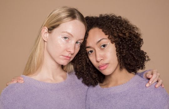 Calm multiracial female friends with colorful makeup wearing similar clothes embracing and looking at camera against beige background in studio