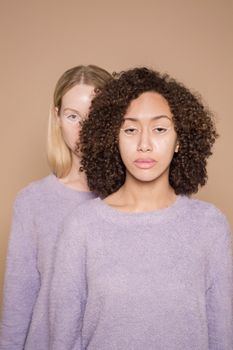 Trendy diverse female friends wearing purple clothes with graphic eyeliner on eyes looking at camera against beige background in studio