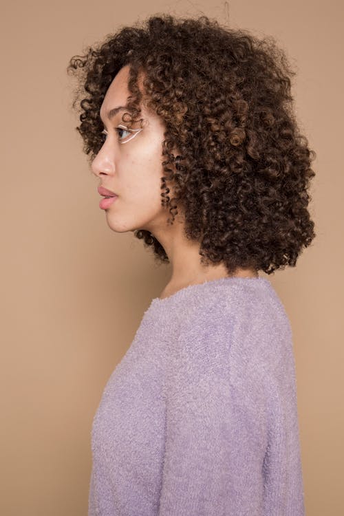 Side view of ethnic female with curly hair and colorful graphic liner wearing purple sweater standing against beige background in studio