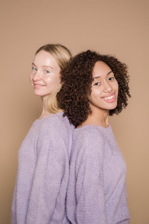 Free Positive multiracial female friends with graphic liner wearing similar clothes looking at camera while standing back to back against beige background Stock Photo