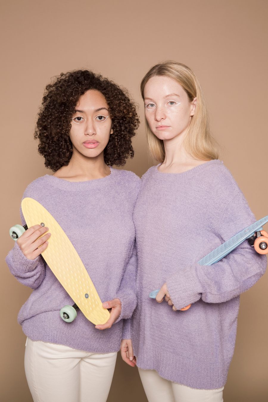 Two friends in lilac knit sweaters holding skateboards against a warm sand backdrop — photo by Michelle Leman on Pexels