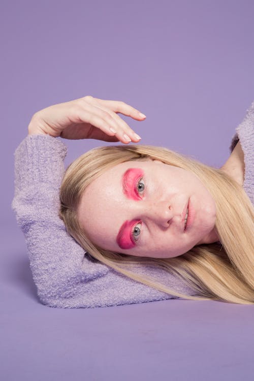 Gentle female with extraordinary makeup looking at camera while lying on floor on purple background of studio