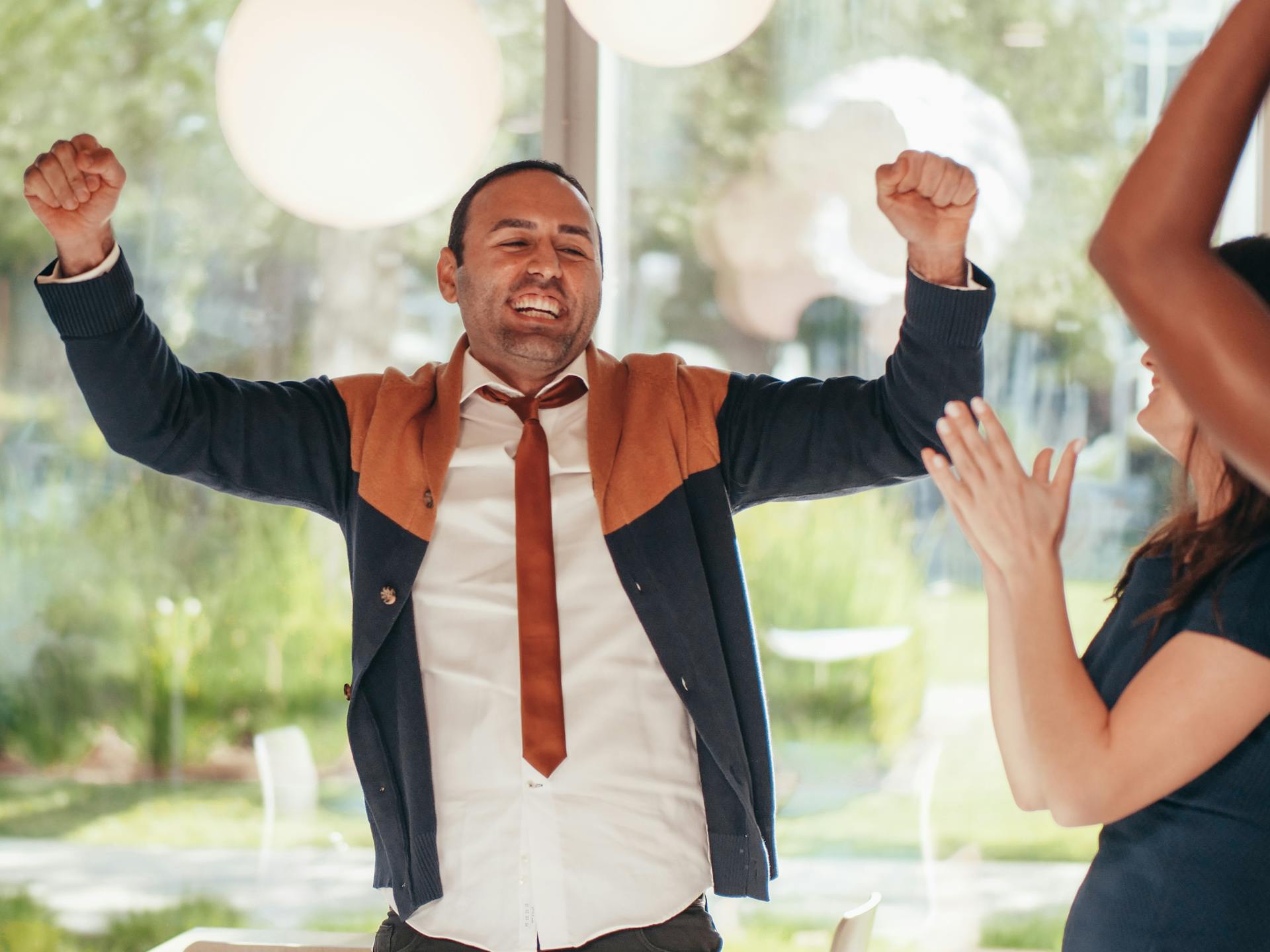 Smiling ethnic male employee in formal apparel with fists up against crop unrecognizable coworkers in workspace