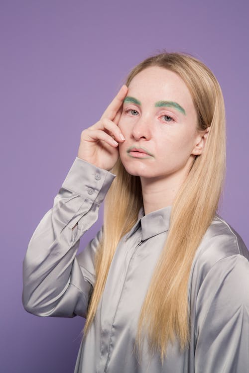Stylish pale female with green eyebrows looking at camera on purple background of studio