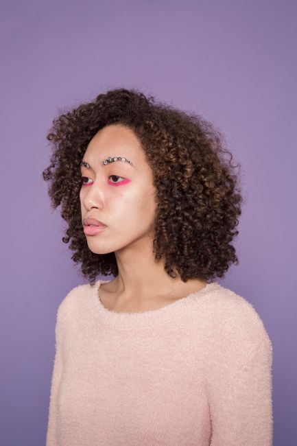 Young serious ethnic lady in sweater and colorful makeup on face with rhinestones on eyebrows looking away in bright studio on violet background