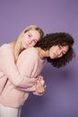 Young serious multiracial girlfriends in sweaters and colorful pink makeup on face looking at camera while hugging in bright studio on violet background