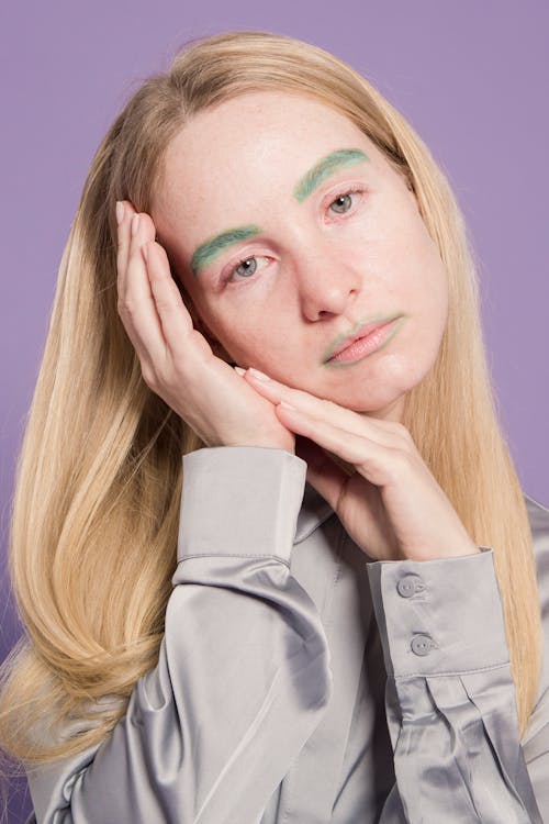 Tired young female in gray shirt with green colorful makeup on face looking at camera on purple background in light studio