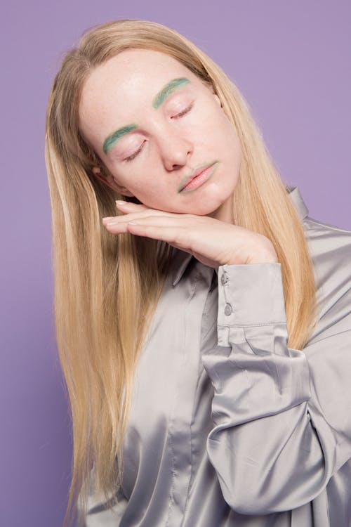 Young sad lady in gray blouse with colorful green dyed eyebrows closing eyes and leaning chin on hand on violet background