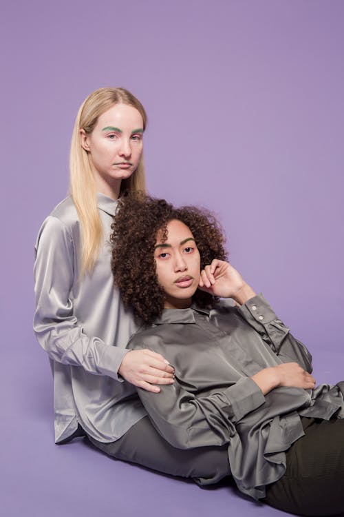 Serious young multiethnic female friends in blouses looking at camera while sitting on floor and leaning on each other on violet background in bright studio