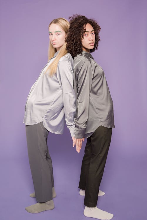 Full body side view of young serious multiracial girlfriends in shirts standing back to back while looking at camera in bright studio on violet background