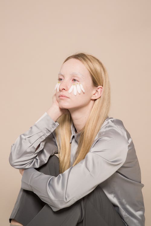 Sad young female in gray shirt with white flower petals on cheeks with hand on face looking away on beige background while sitting in light studio