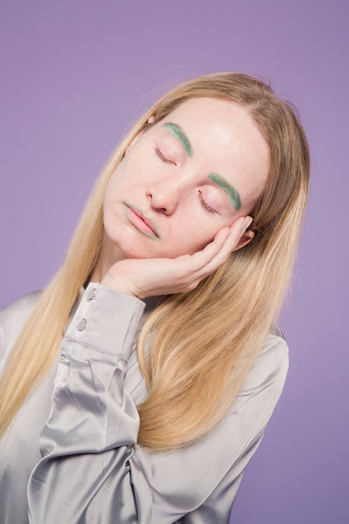 Tired young female in gray shirt with green colorful makeup on face with hand on face and closed eyes on purple background in light studio
