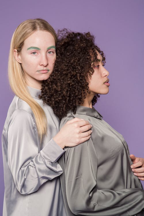 Serious young multiethnic female friends in blouses standing close to each other on violet background in bright studio