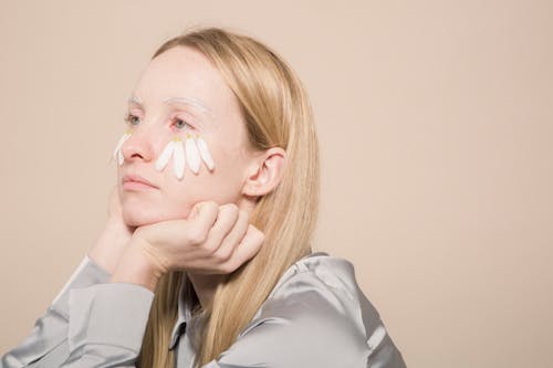 Calm female with with organic chamomile petals on face for natural skincare looking away against beige background in modern studio