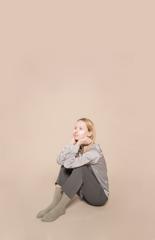 Full body of young female with organic white petals on face for skincare sitting against beige background in modern studio