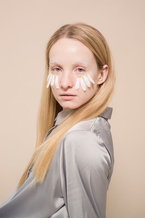 Side view of self assured young female model with long blond hair and chamomile petals on face looking at camera against beige background