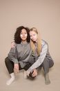Full length of stylish young multiethnic female models with petals of fresh flowers on faces sitting on haunches and cuddling against beige background