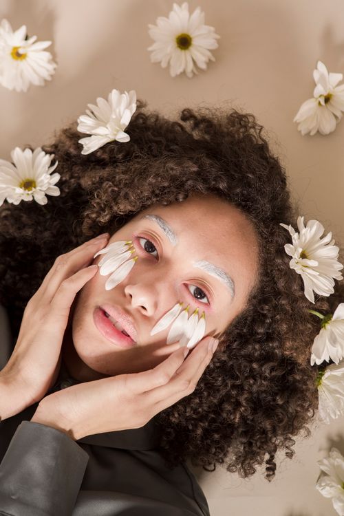 Free From above of calm young ethnic female model touching cheeks and looking at camera while lying with fresh chamomiles in curly hair and petals on face Stock Photo