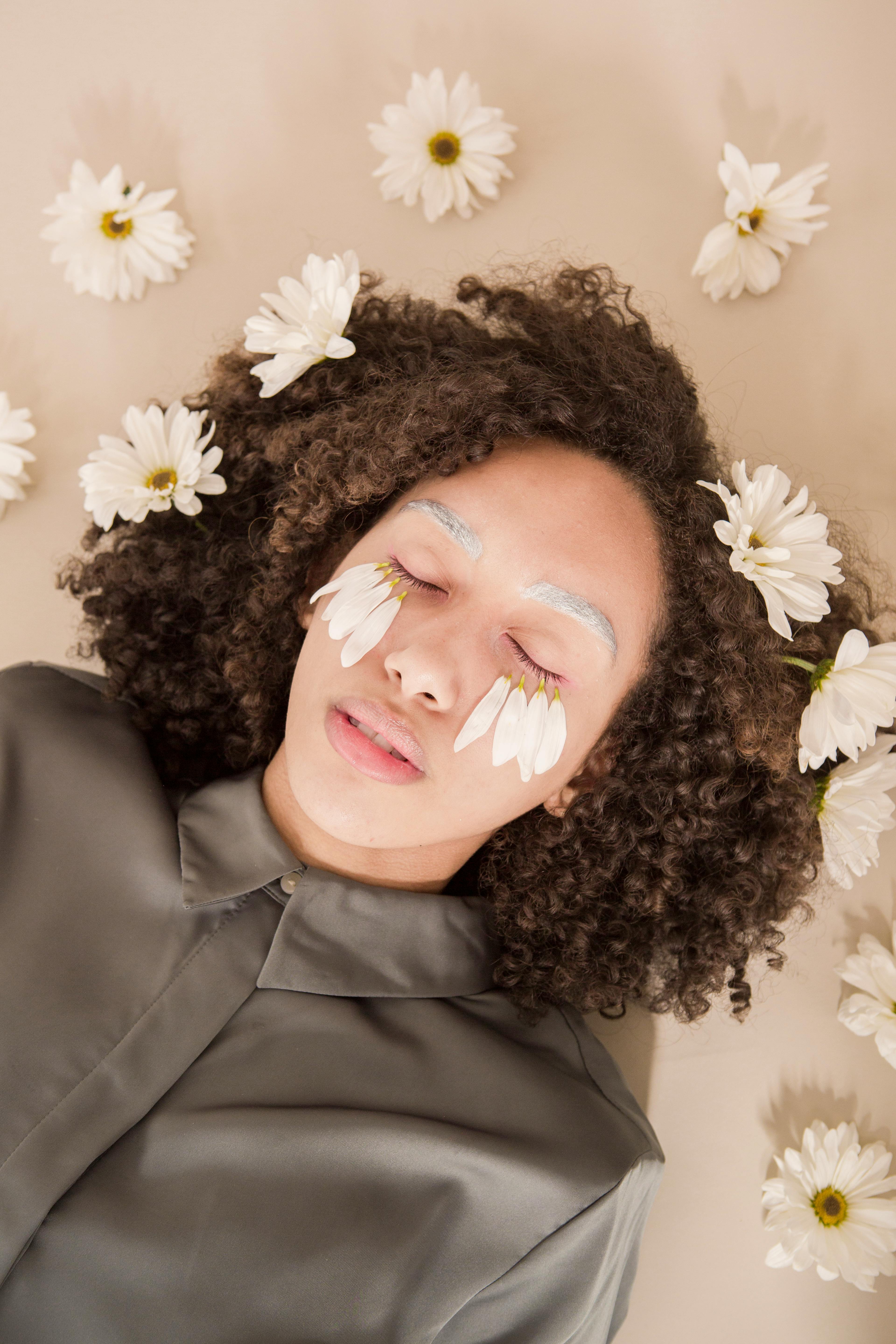 Top view of relaxed young ethnic lady with dark curly hair lying in beige surface with closed eyes and flower petals on face near scattered chamomiles