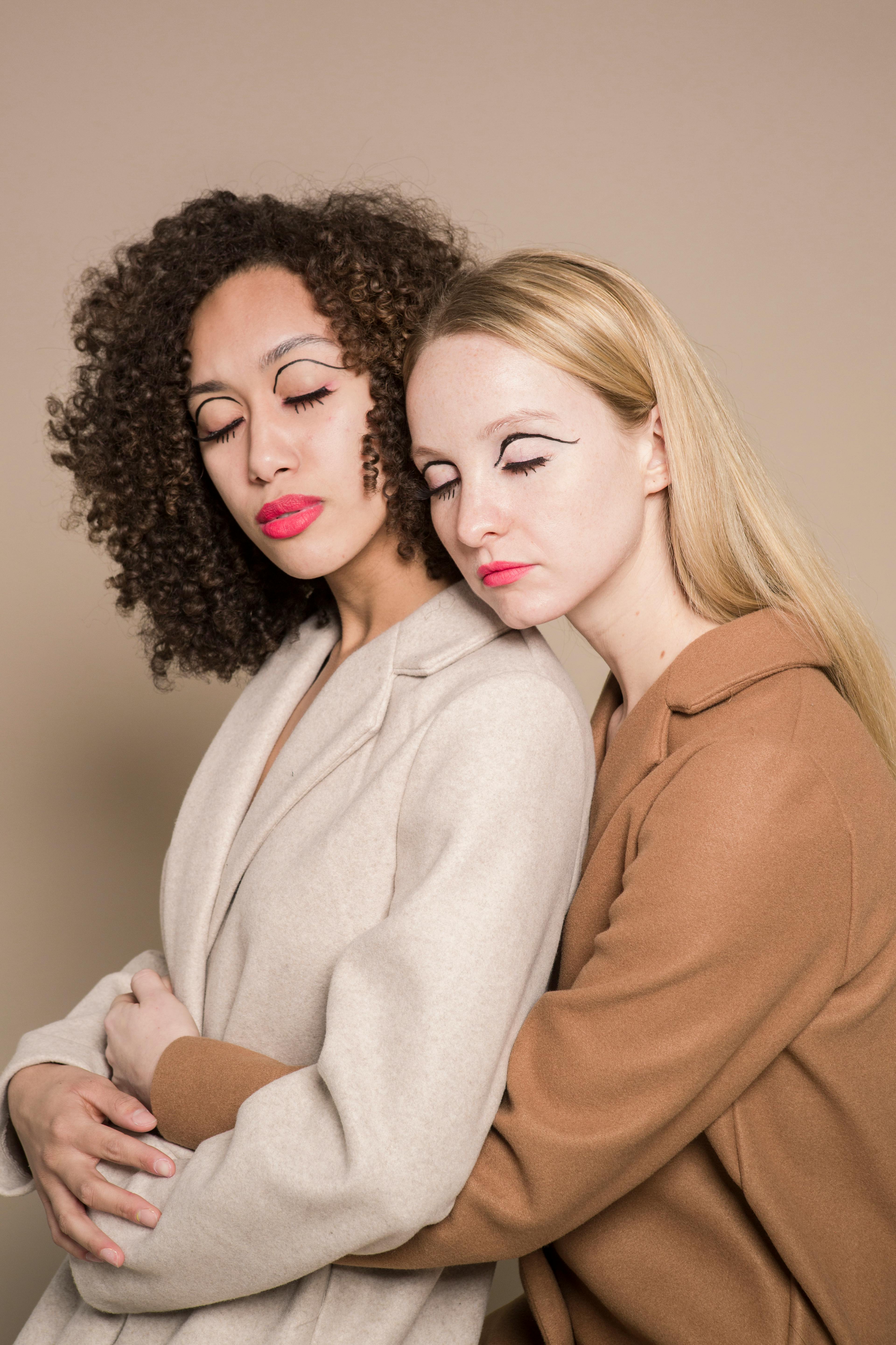 Multiethnic extraordinary friends with bright makeup in stylish coats hugging on beige background of studio